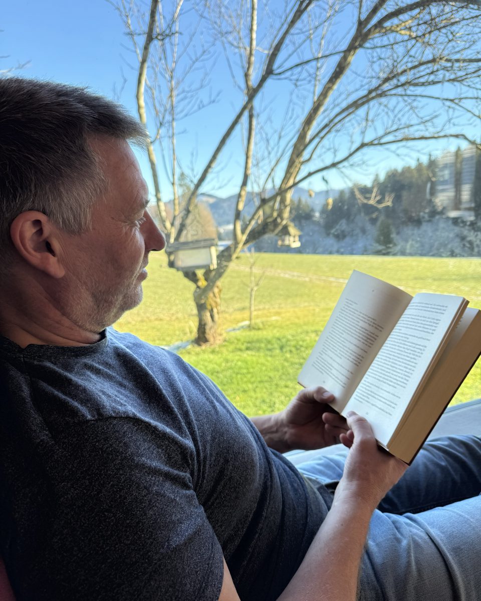 Titus J. Rinke liest in einem Buch, im Hintergrund Wiese und Bäume im Allgäu
